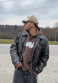 Muž s tetovaním na krku, ktorý nosí maskáčovú čapicu, koženú bundu, hnedý mikinu a džínsy, stojí na vydláždenej ploche a pozer�á doľava, s stromami a zamračenou oblohou v pozadí.