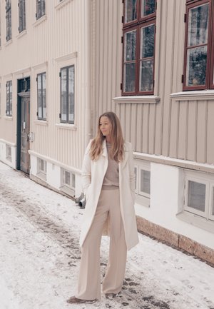 Mujer con pantalones beige, suéter y abrigo blanco de pie en una calle nevada junto a un edificio de madera beige con ventanas enmarcadas en rojo.