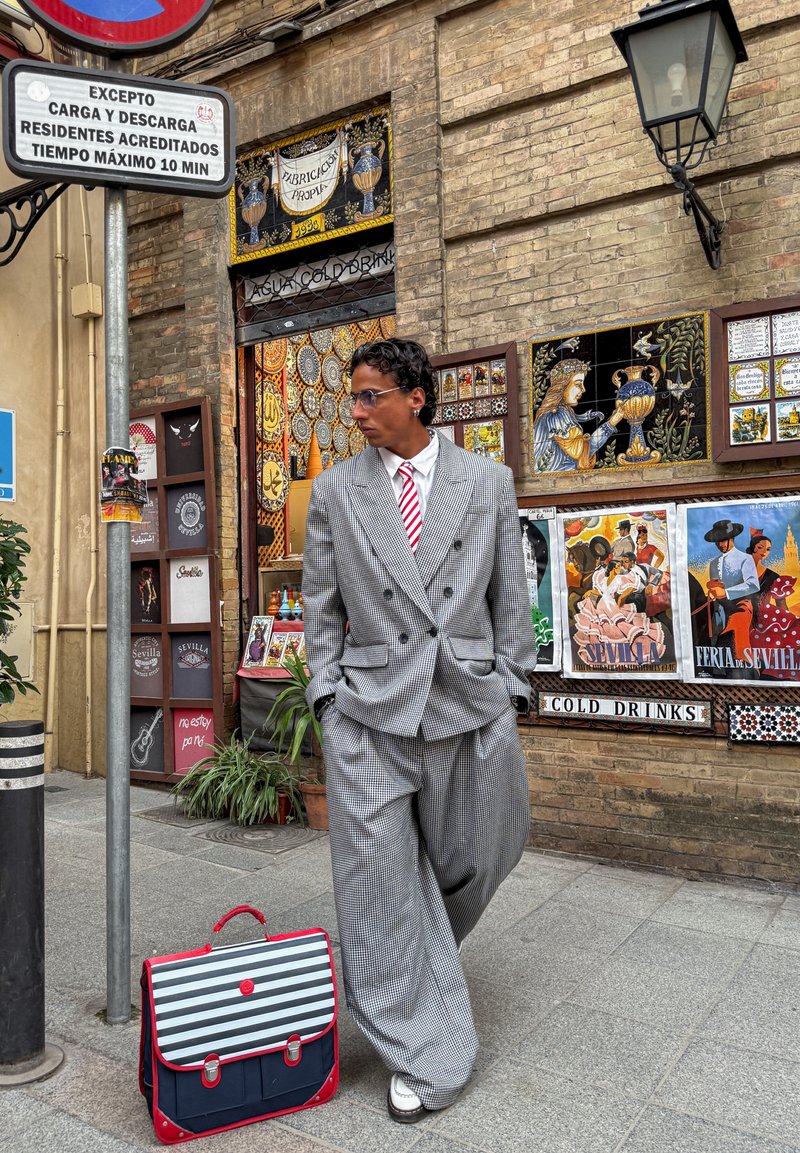 Homme en costume gris à carreaux surdimensionné et cravate rouge rayée debout à côté d'une mallette rayée dans une rue avec un mur de briques décoré et des affiches.
