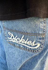 Blå denimbukser med en baklomme med hvit brodert "Dickies"-logo, dekorert med oransje søm.