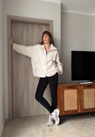 Mujer con chaqueta de forro polar blanca, pantalones negros y zapatillas, de pie dentro de una habitación apoyada en el marco de una puerta de madera junto a un mueble de televisión de madera.