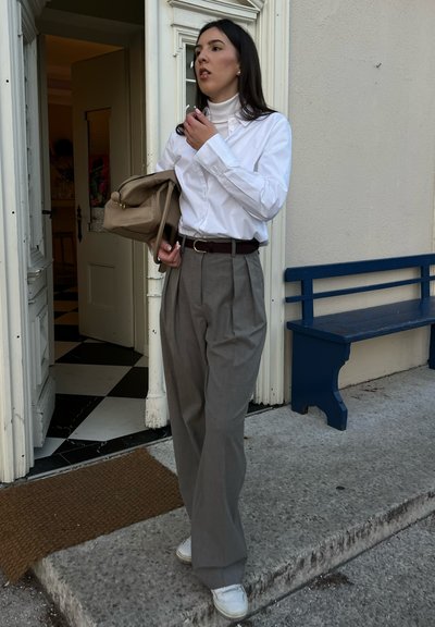 Camisa blanca de botones, pantalones gris de cintura alta con pliegues, cinturón marrón, sosteniendo un bolso color beige, zapatillas blancas, de pie cerca de una puerta.