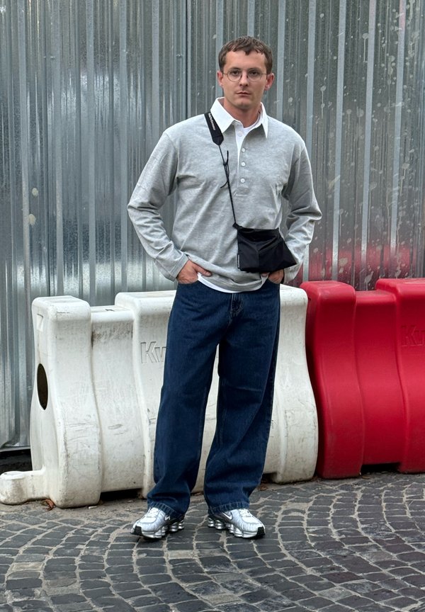 Homem com óculos, vestindo uma camisa cinza de manga longa, jeans escuros, ténis prateados e uma mochila cruzada preta, está em pé sobre paralelepípedos perto de barreiras vermelhas e brancas.