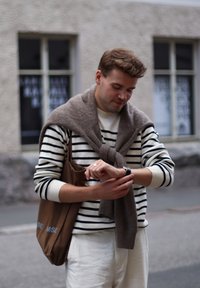 Striped jumper with a light grey wrap, cream trousers, and a brown tote bag. Man checking his watch. Soft textures and neutral tones.