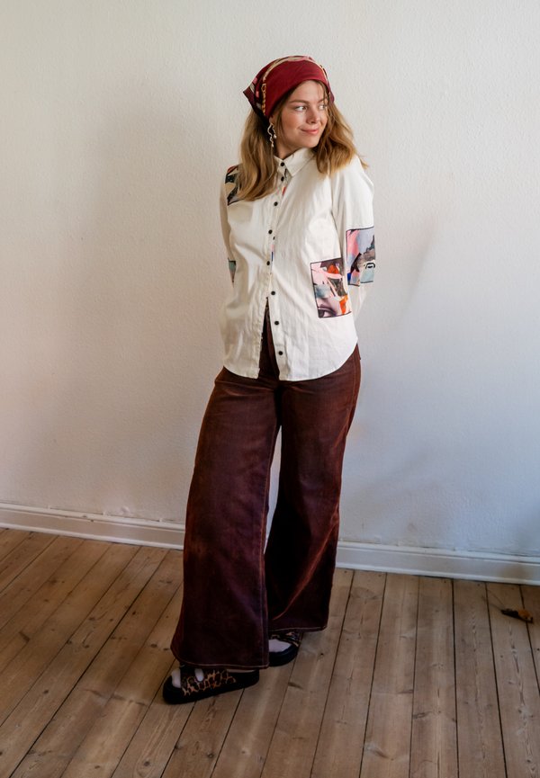 Femme portant une chemise blanche boutonnée avec des patches colorés, un pantalon marron large, des chaussures à motif léopard et un foulard rouge sur la tête, debout sur un sol en bois.