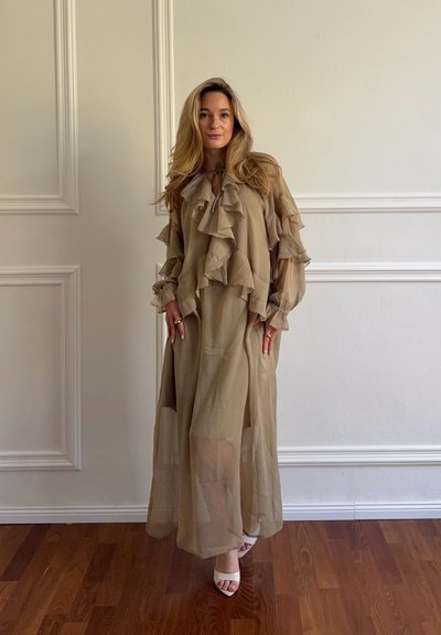 Mujer con cabello largo y rubio, vestida con un vestido beige transparente con volantes y tacones blancos abiertos, de pie sobre un suelo de madera frente a paneles blancos en la pared.