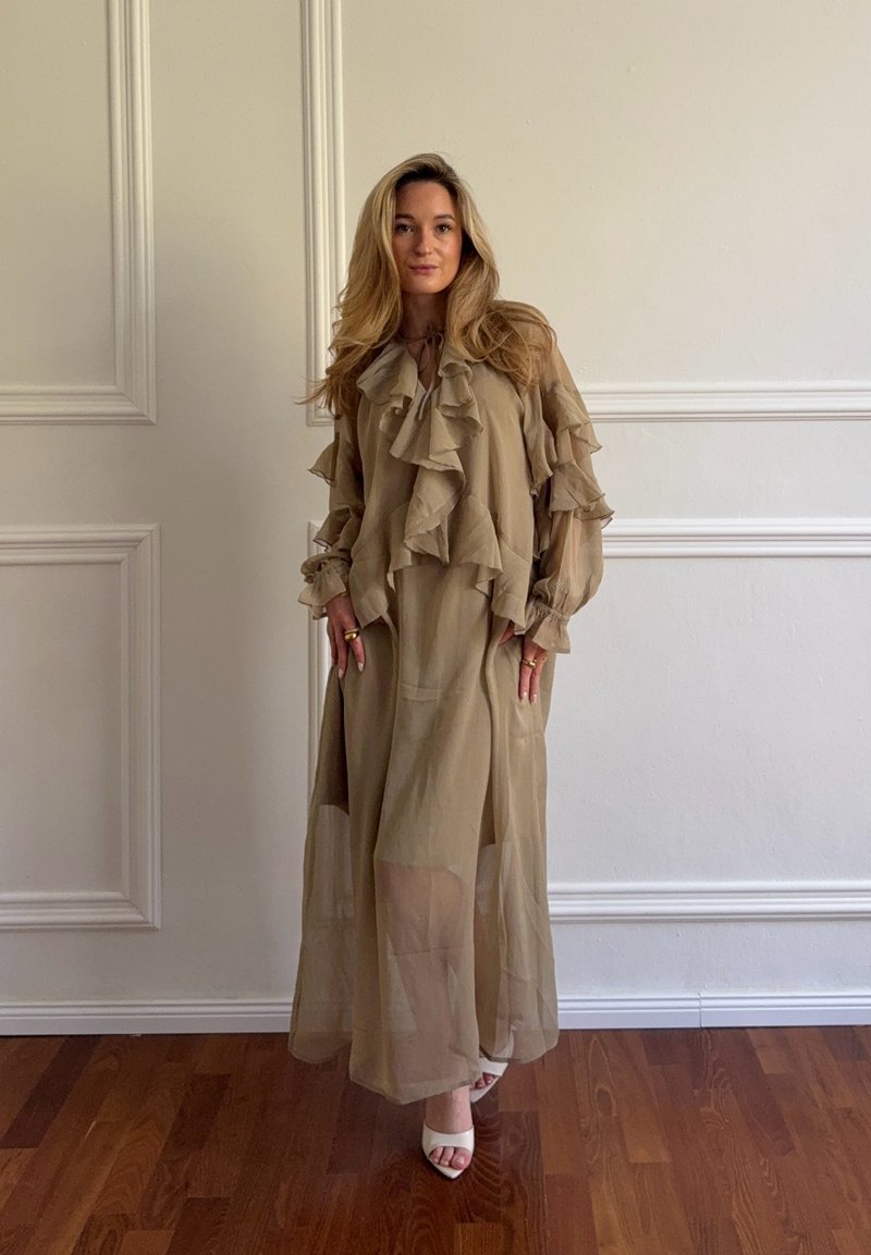 Femme aux longs cheveux blonds portant une robe beige transparente à volants et des talons blancs ouverts, debout sur un sol en bois devant des panneaux muraux blancs.