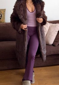 Personne portant un long manteau en fourrure brun foncé sur un pantalon violet ajusté et un débardeur lavande, debout sur un sol en bois à côté d'un canapé marron.