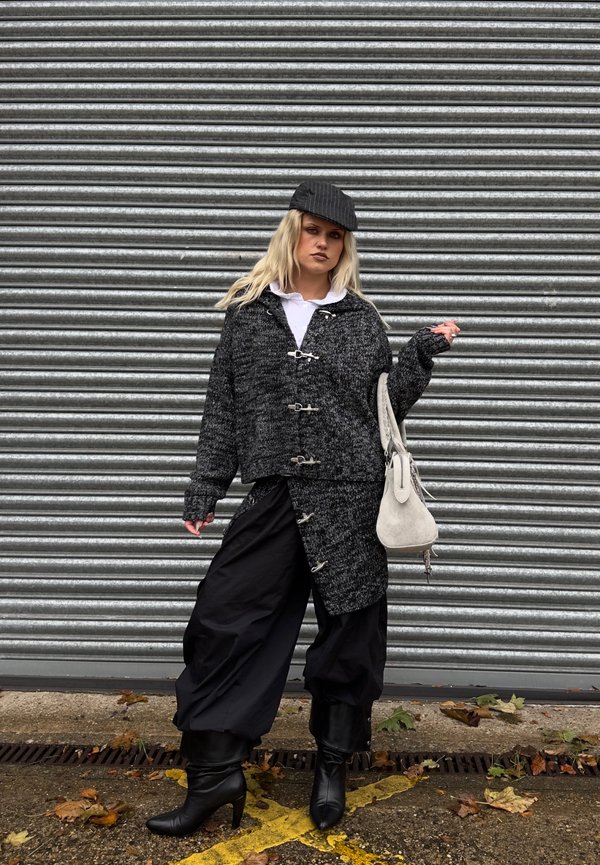 Une femme pose devant un volet métallique, vêtue d’un ensemble sombre en tweed, d’une casquette noire et de bottes, et portant un sac à main clair.