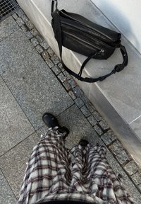 Crna kožna torba preko tijela s više odvojenih pretinaca na zip; stilizirana s kariranim, laganim, lepršavim hlačama i crnim gležnjačama.