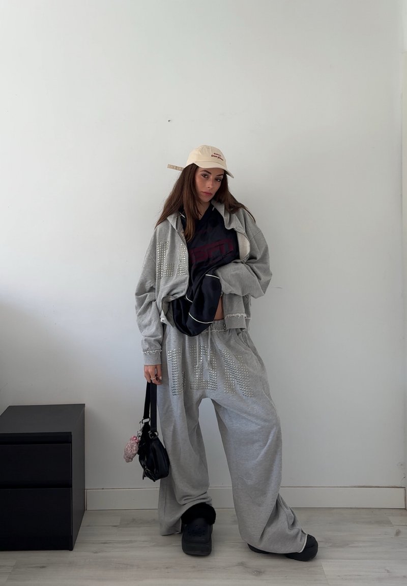 Jeune femme portant un sweat à capuche gris oversize et un pantalon avec des strass, un top noir, une casquette beige, un sac à main noir, debout contre un mur blanc.