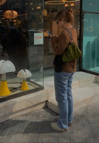 Uma mulher com um suéter castanho de malha e jeans azuis está em frente a uma loja, olhando para lâmpadas brancas e amarelas na vitrine.