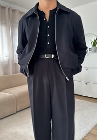 En person iført en svart skjorte med knapper, en ujevn svart jakke, svart belte og høyhalsede svarte bukser står innendørs med hendene i lommer.