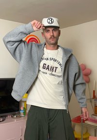 Szara bluza na suwak nałożona na białą koszulkę z napisem "GANT". Luźne, oliwkowozielone szorty oraz biała czapka z daszkiem z numerem 3.