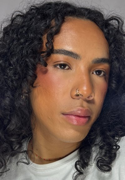 Persona con cabello negro rizado, ojos marrones, piercing en la nariz y maquillaje natural, que lleva una camiseta blanca y un delicado collar de cuentas.