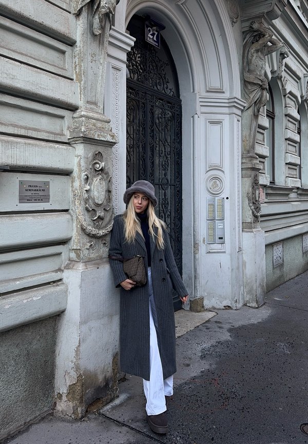 Soprabito grigio a righe sottili, pantaloni bianchi, cappello grigio; in piedi davanti a un edificio con una texture ornata e un cancello in ferro battuto. Borsa marrone.