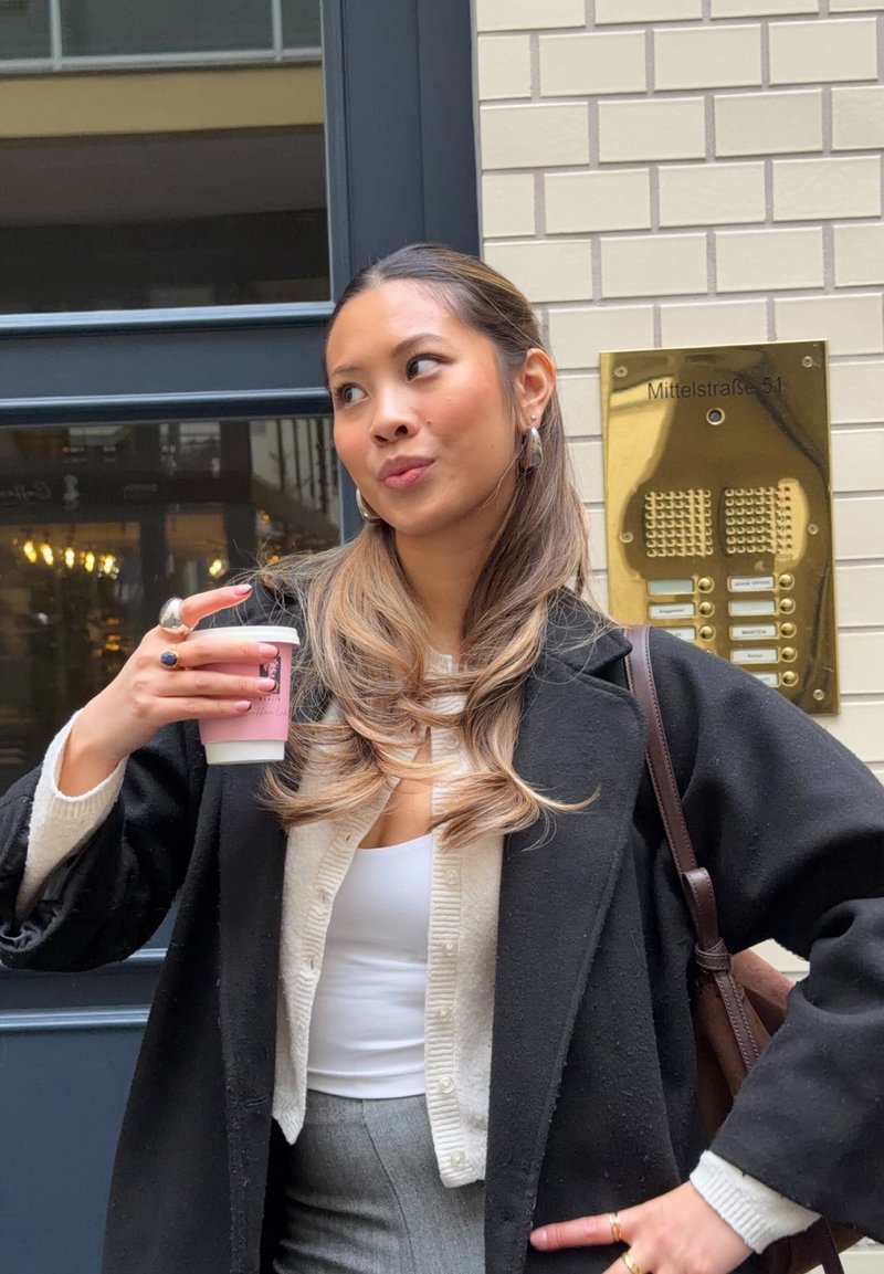 Una donna tiene una tazza di caffè rosa, indossando un cappotto nero oversize sopra un maglione crema con bottoni e pantaloni grigi. Interfono a parete visibile.