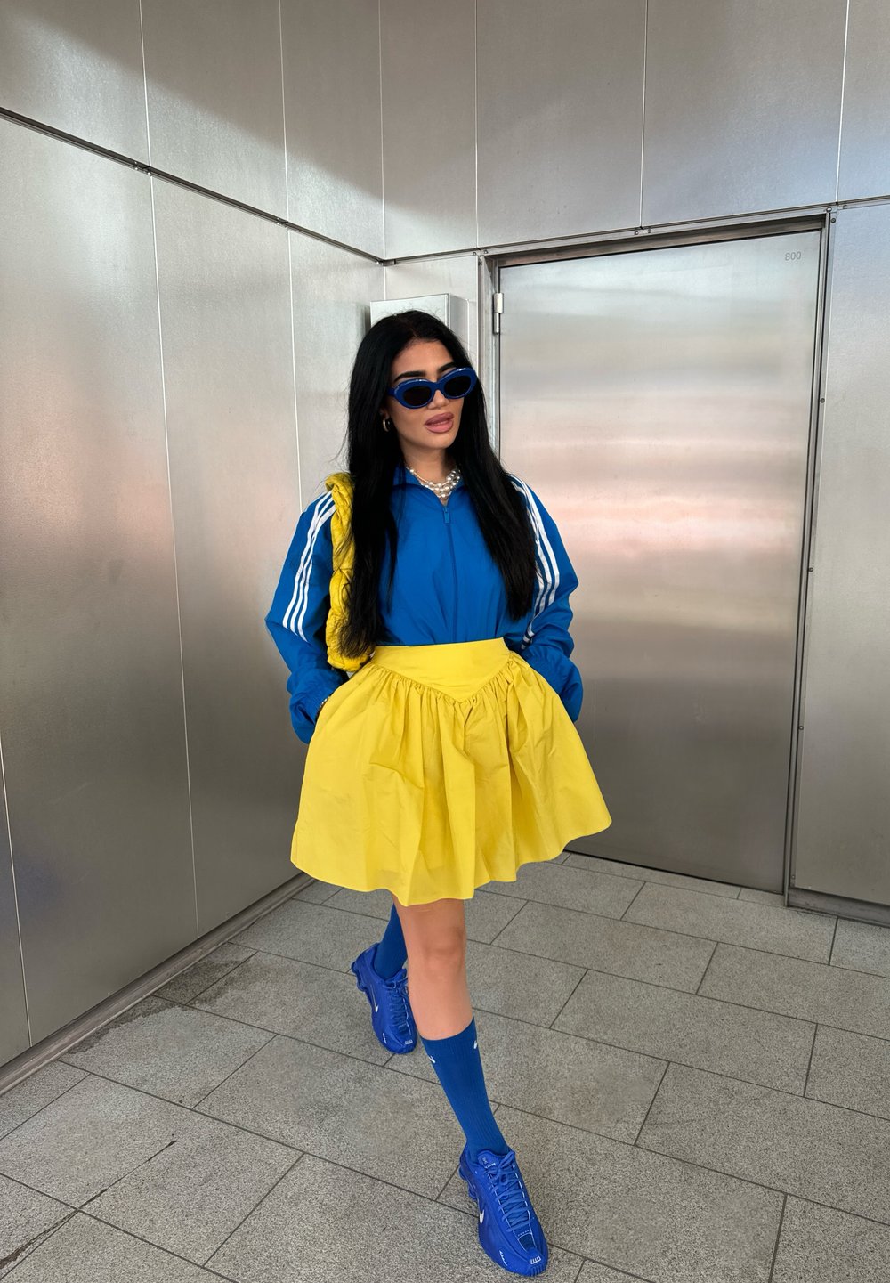 Giacca sportiva blu con strisce bianche, gonna gialla, occhiali da sole blu, calzini blu e scarpe blu. Texture lisce, colori vivaci.
