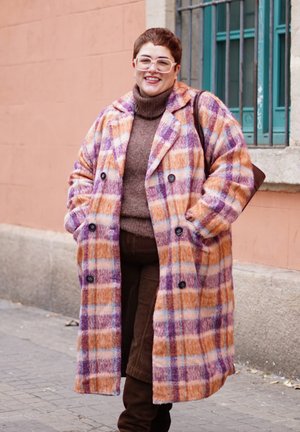 Mujer sonriendo y de pie en una calle, vistiendo un abrigo a cuadros colorido, un suéter de cuello alto marrón, pantalones marrones y gafas con montura blanca.