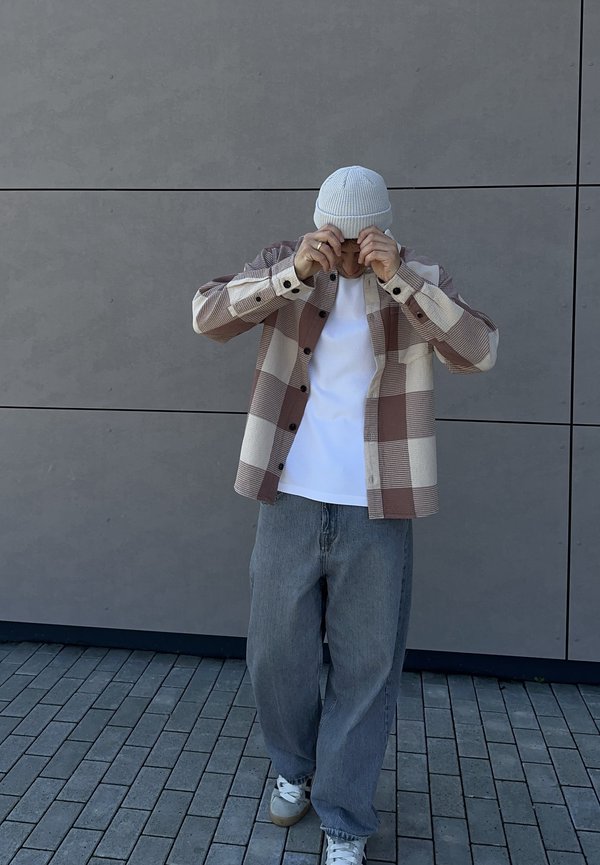 Person, die weiße Strickmütze richtet, trägt eine übergroße beige-braun karierte Jacke, weißes Hemd, lockere graue Jeans und weiße Sneakers und steht auf gefliestem Boden.