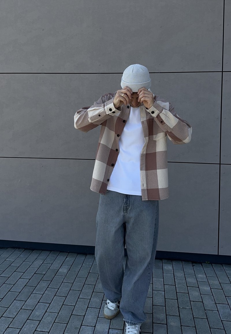Personne ajustant un bonnet en tricot blanc, portant une veste oversize beige et marron à carreaux, une chemise blanche, un jean gris ample et des baskets blanches, debout sur un sol carrelé.