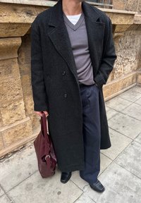 Άτομο που φοράει μακρύ σκούρο παλτό, γκρι πουλόβερ με V στο λαιμό πάνω από λευκό πουκάμισο, παντελόνι με λεπτές ρίγες, μαύρα παπούτσια, κρατώντας μια μπορντό δερμάτινη τσάντα σε πεζοδρόμιο στην πόλη.