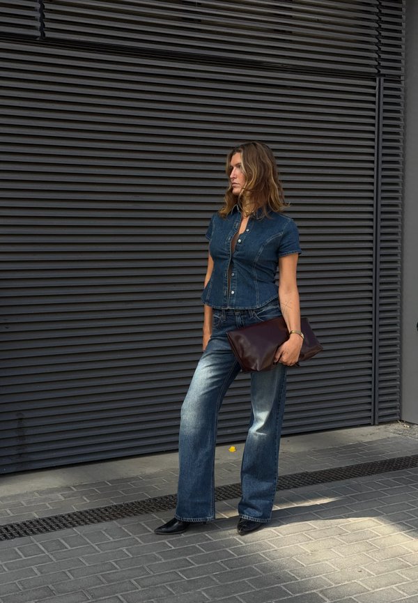 Femme portant une chemise en denim et un jean tient une grande pochette sombre, debout contre un mur noir à lamelles horizontales dans une rue pavée.
