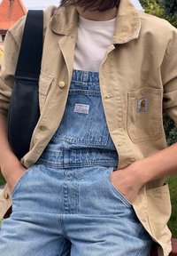 Άτομο με μπεζ τζάκετ Carhartt πάνω από λευκό πουκάμισο και τζιν σαλβάρι Levi's, μαύρη τσάντα ώμου, χέρια στις τσέπες, σε εξωτερικό χώρο με θολό φόντο.