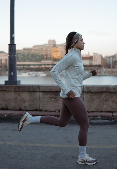 Camiseta de manga larga gris claro para correr, mallas ajustadas marrones, calcetines deportivos blancos y zapatillas de correr de color claro; niña en movimiento junto a un paseo marítimo.
