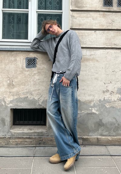 Jersey gris de punto, jeans de mezclilla azul sueltos con cintura elástica, botas beige tipo slip-on, de pie contra una pared texturizada.