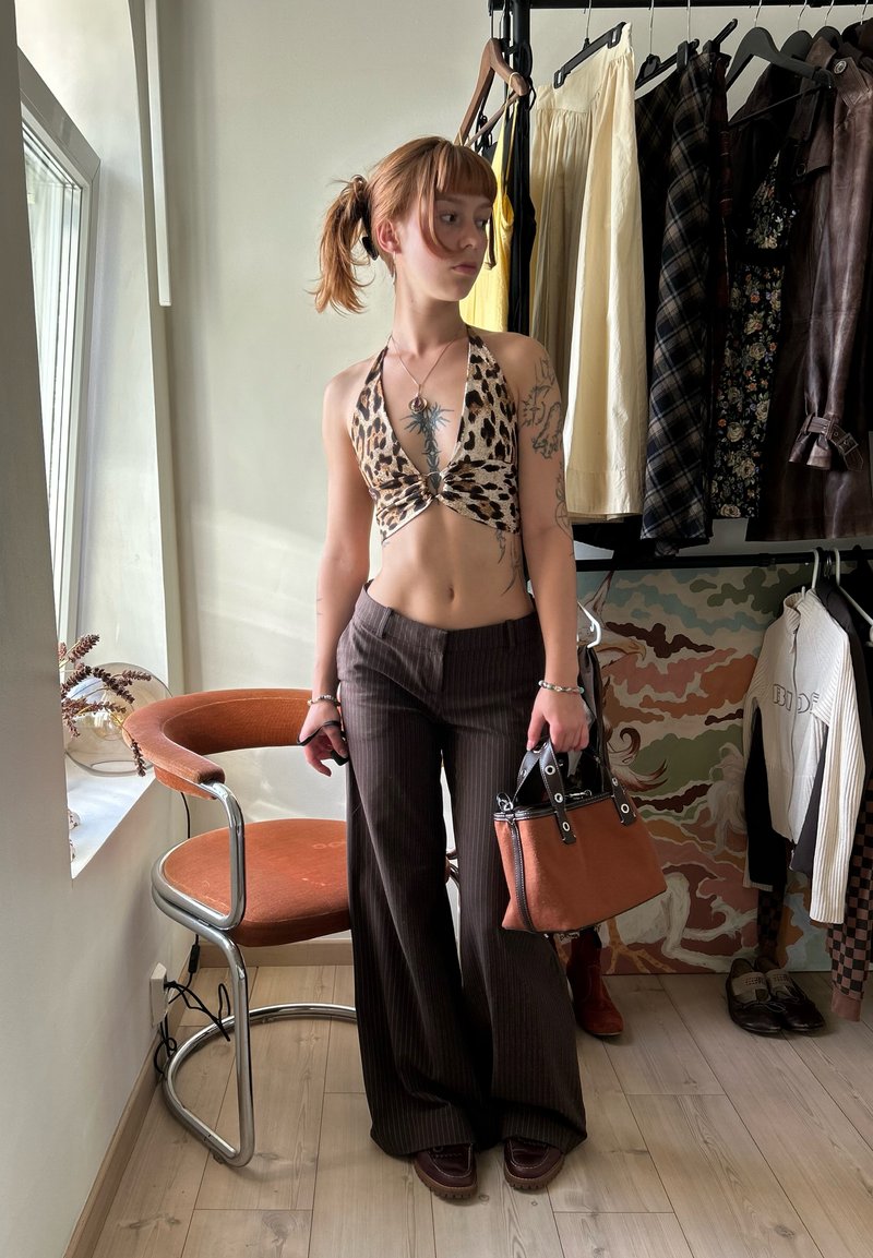 Jeune personne tatouée portant un haut dos nu à imprimé léopard et un pantalon marron, tenant un sac à main marron dans une pièce avec des portants à vêtements et une chaise.