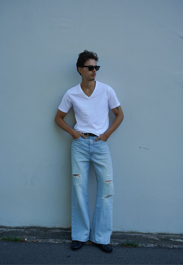 Homme portant des lunettes de soleil noires, un t-shirt blanc à col en V, des jeans larges bleu clair déchirés et des chaussures noires, debout contre un mur gris.