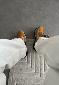 Bottes de randonnée marron et beige portées avec un pantalon blanc, posées sur un tapis de sol texturé gris clair avec un fond de sol gris.