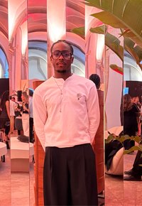 Fehér hosszú ujjú ing virágmintával, fekete bőszárú nadrág és kerek szemüveg. Tükrök és zöld növények egy stílusos belső térben.