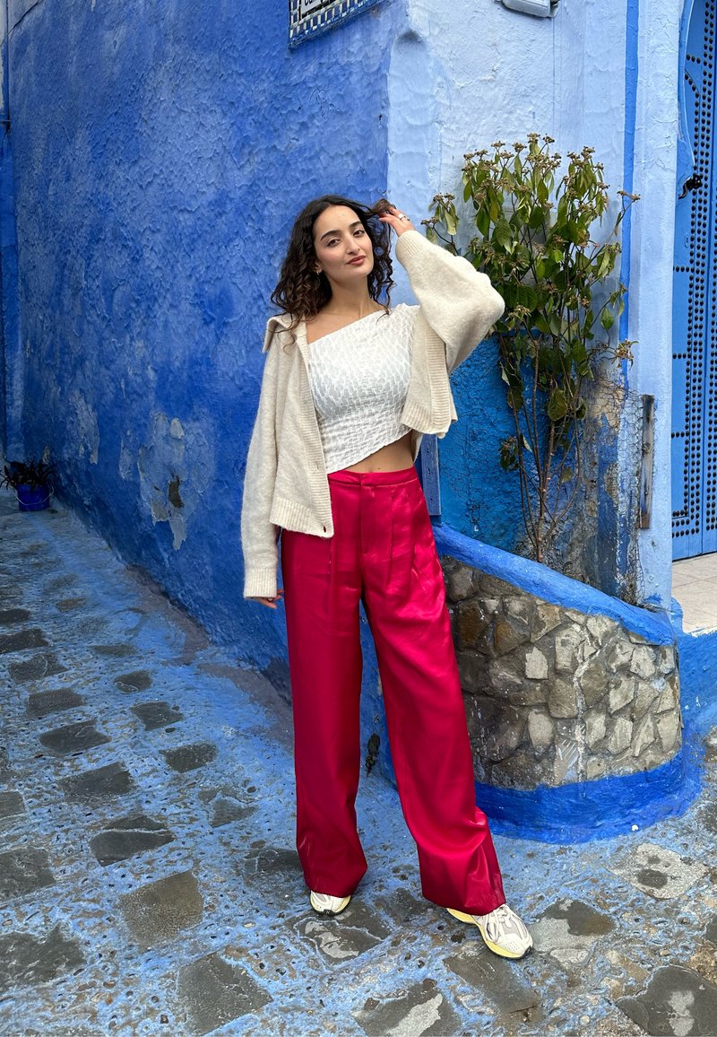 Μοντέλο φορώντας ένα κοντό λευκό υφασμάτινο μπλουζάκι, ένα υπερμεγέθη κρεμ ζακέτα και ψηλόμεσα ροζ παντελόνια με γυαλάδα, ενώ στέκεται δίπλα σε ένα μπλε τοίχο.