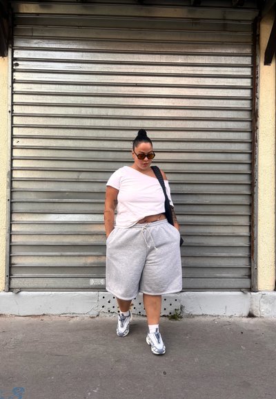 Camiseta rosa claro sin mangas, pantalones cortos grises y zapatillas deportivas blancas con detalles en plata. La persona está de pie contra una persiana metálica enrollable.
