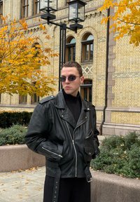 Svart läderbikerjacka med silverdetaljer, figurnära design och fickor med dragkedja. Bärs över en polotröja, mot en bakgrund av höstlöv.