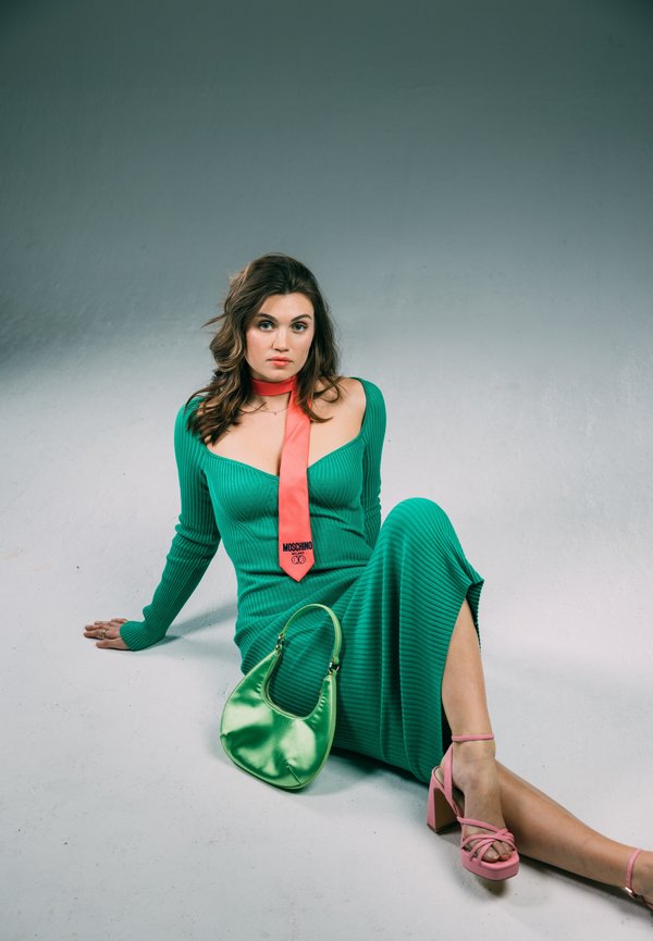 Green ribbed dress with long sleeves, featuring a deep neckline and side slit. Pink strappy heels and a shiny green handbag. Orange Moschino tie accent.