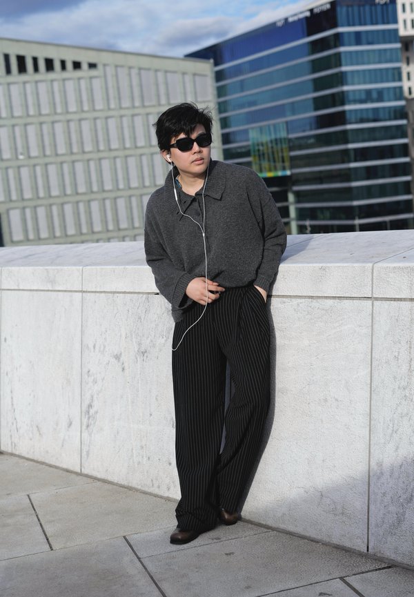 Persona che indossa occhiali da sole scuri, maglione grigio e pantaloni a righe, appoggiata a un muro di pietra bianco con cuffie in un ambiente urbano.