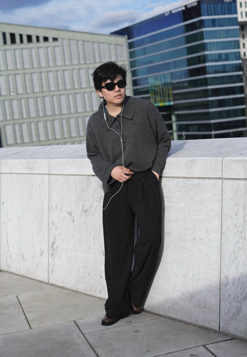 Personne portant des lunettes de soleil foncées, un pull gris et un pantalon rayé, appuyée contre un mur en pierre blanche avec des écouteurs dans un cadre urbain.