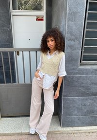 Maglione corto lavorato a maglia beige indossato sopra una camicia bianca, abbinato a pantaloni a gamba larga a righe chiare e sneakers bianche, in piedi davanti a un muro grigio.