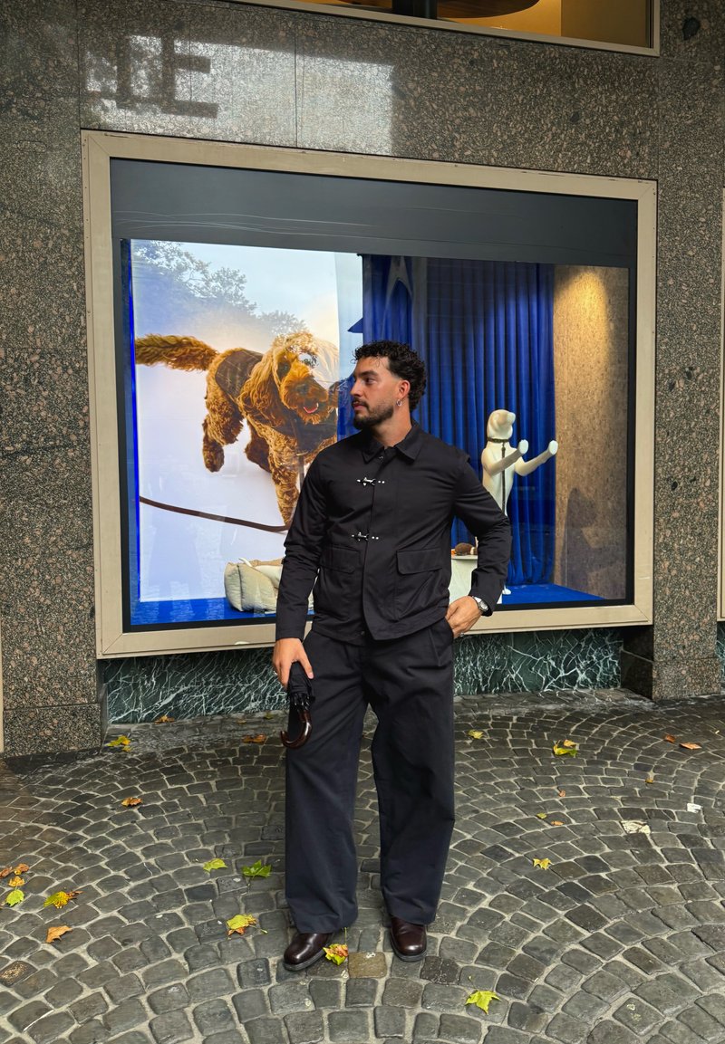 Black button-up jacket with two front pockets, paired with loose black trousers and brown leather shoes. Umbrella held in the right hand.