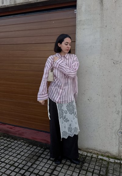 Mujer con cabello corto y oscuro, vestida con camiseta de rayas rosa, pantalones negros con encaje blanco encima, sosteniendo un pequeño bolso tejido, parada junto a una puerta de garaje.