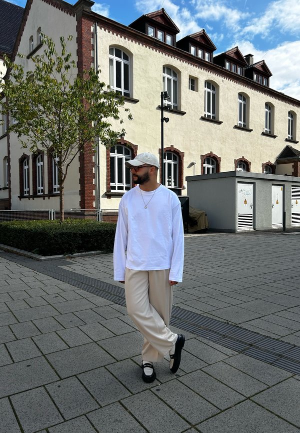 Camisa blanca de manga larga, pantalones beige de pierna ancha y zapatos negros con acentos blancos. De pie en un área pavimentada cerca de un edificio con grandes ventanales.