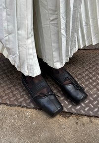 Zwarte leren platte schoenen met vierkante neus, veters gestrikt, gedragen met doorzichtige zwarte sokken met stippen. Witte geplooide rok zichtbaar erboven. Metalen vloer eronder.