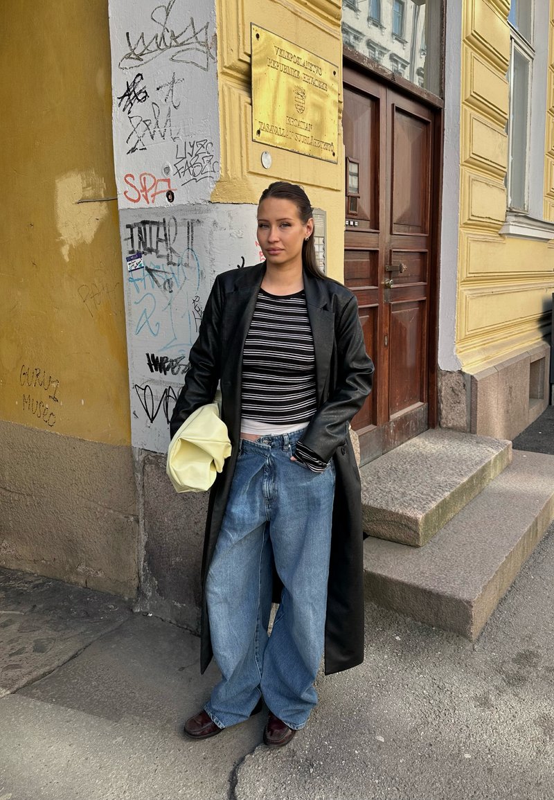 Young woman in long black coat and striped shirt stands on city sidewalk near graffiti-covered wall and wooden door holding yellow bag.