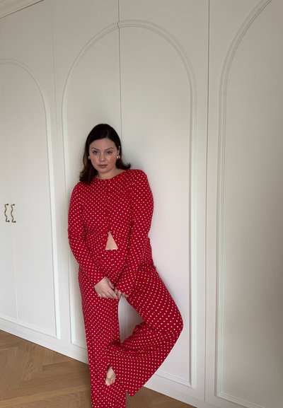Mujer apoyada contra un armario blanco, vestida con un conjunto de top y pantalón de manga larga rojo con lunares, posando con una pierna doblada y las manos entrelazadas.