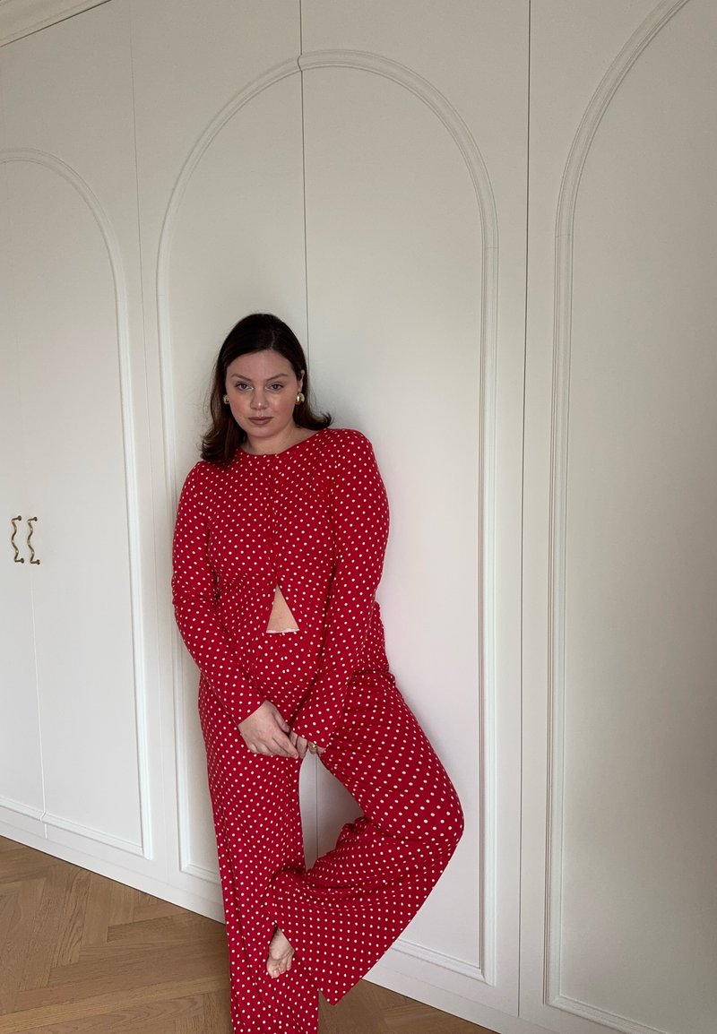 Mujer apoyada contra un armario blanco, vestida con un conjunto de top y pantalón de manga larga rojo con lunares, posando con una pierna doblada y las manos entrelazadas.