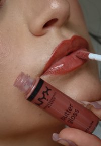 NYX Butter Gloss in einer klaren Tube mit schwarzem Etikett, auf vollen Lippen aufgetragen, zeigt einen glänzenden, warmen Nude-Ton. Weiche und glatte Textur.