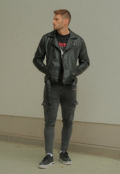 Chaqueta de cuero negro estilo biker con cierre, combinada con pantalones cargo negros que tienen bolsillos laterales. Zapatillas negras con suelas blancas. Fondo neutro.